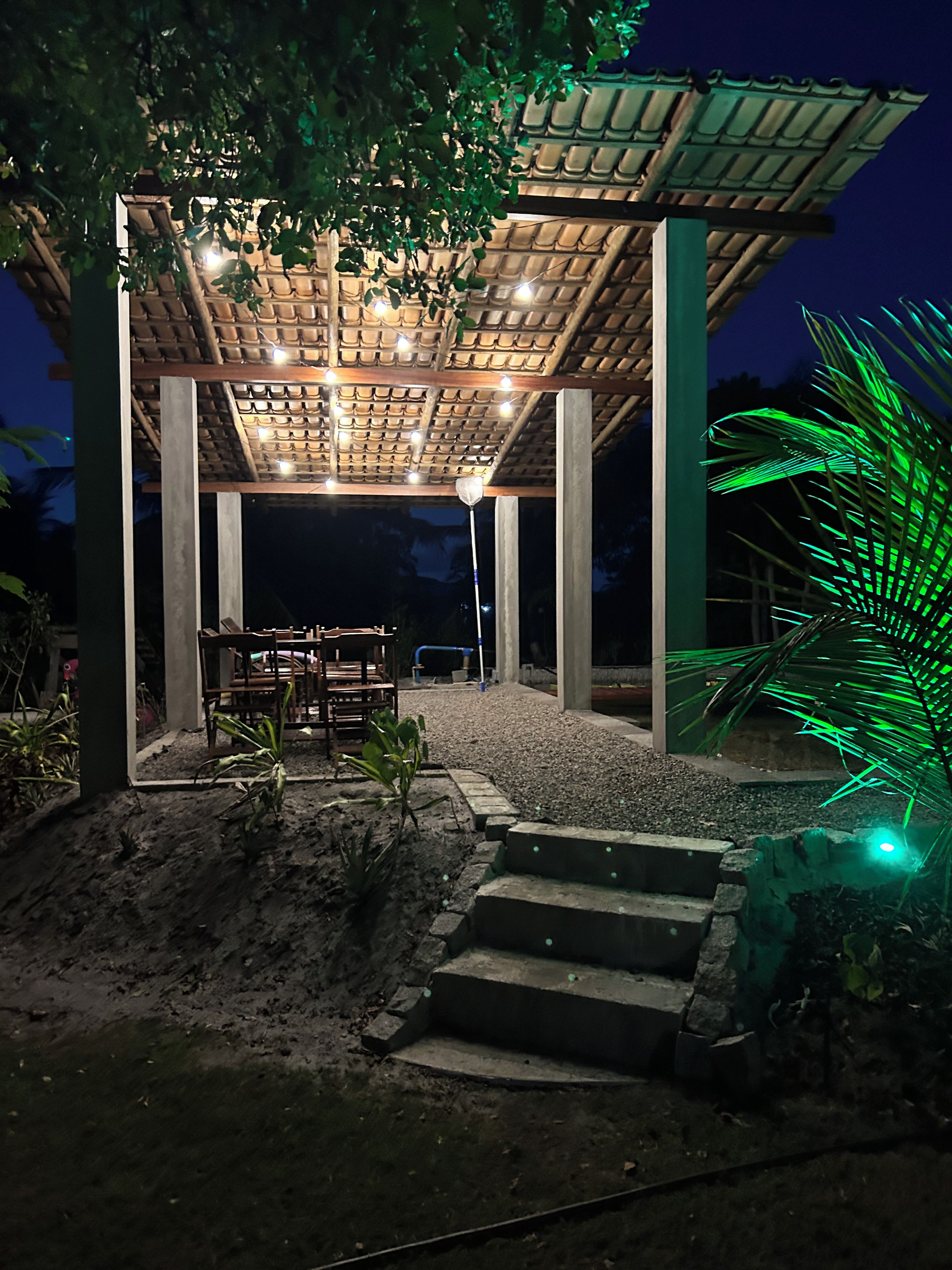 Un espacio de estar al aire libre techado, con una mesa y sillas, rodeado de un jardín con una palmera.