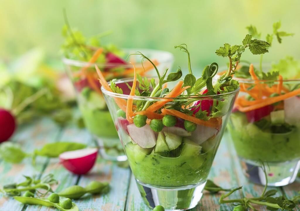 A clear glass with a layered salad, including green peas, carrots, and radishes, on a wooden surface.