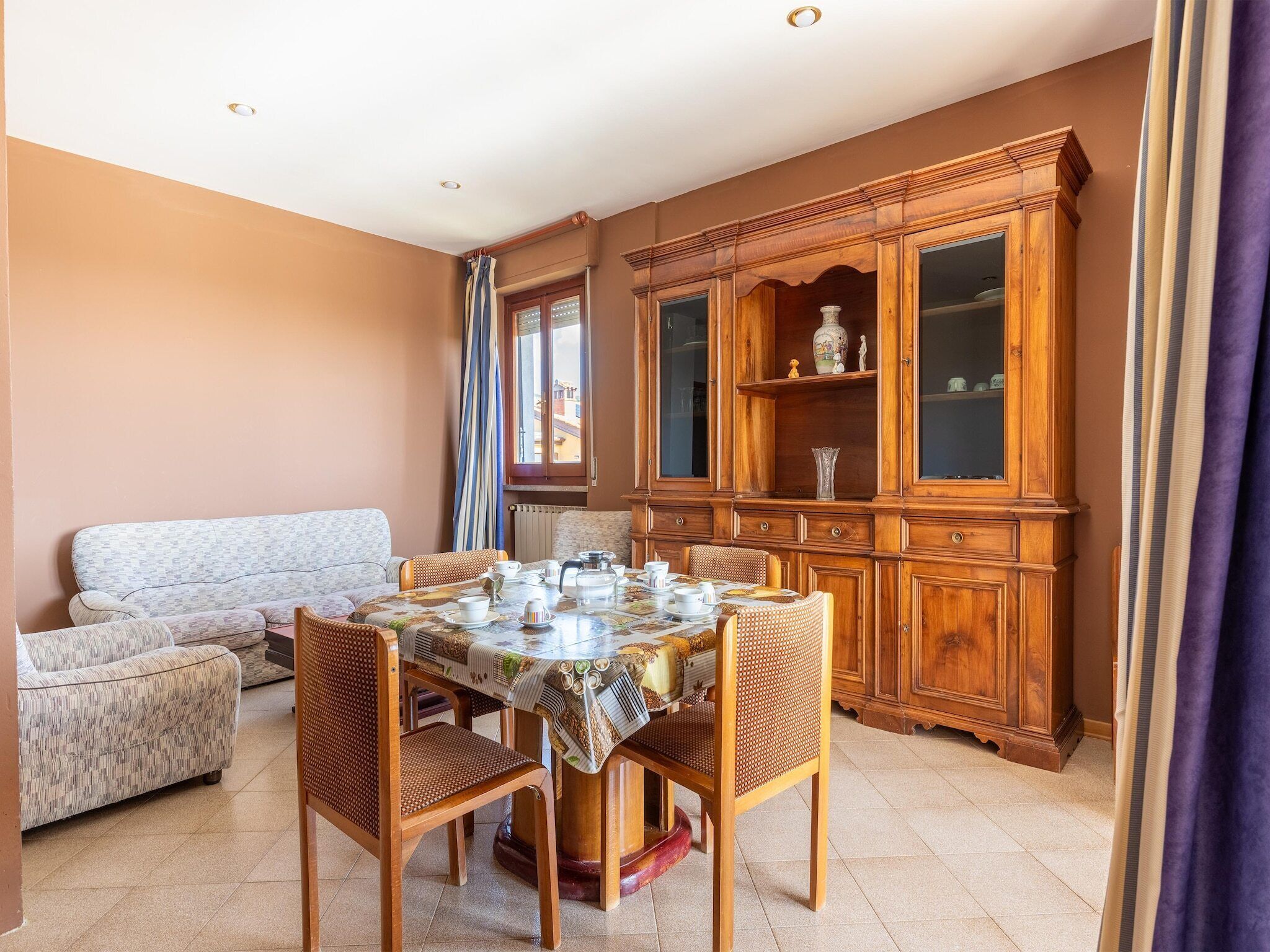 Un comedor con una mesa redonda preparada para cuatro personas, rodeada de sillas de mimbre. Un aparador de madera con puertas de cristal está apoyado contra la pared.