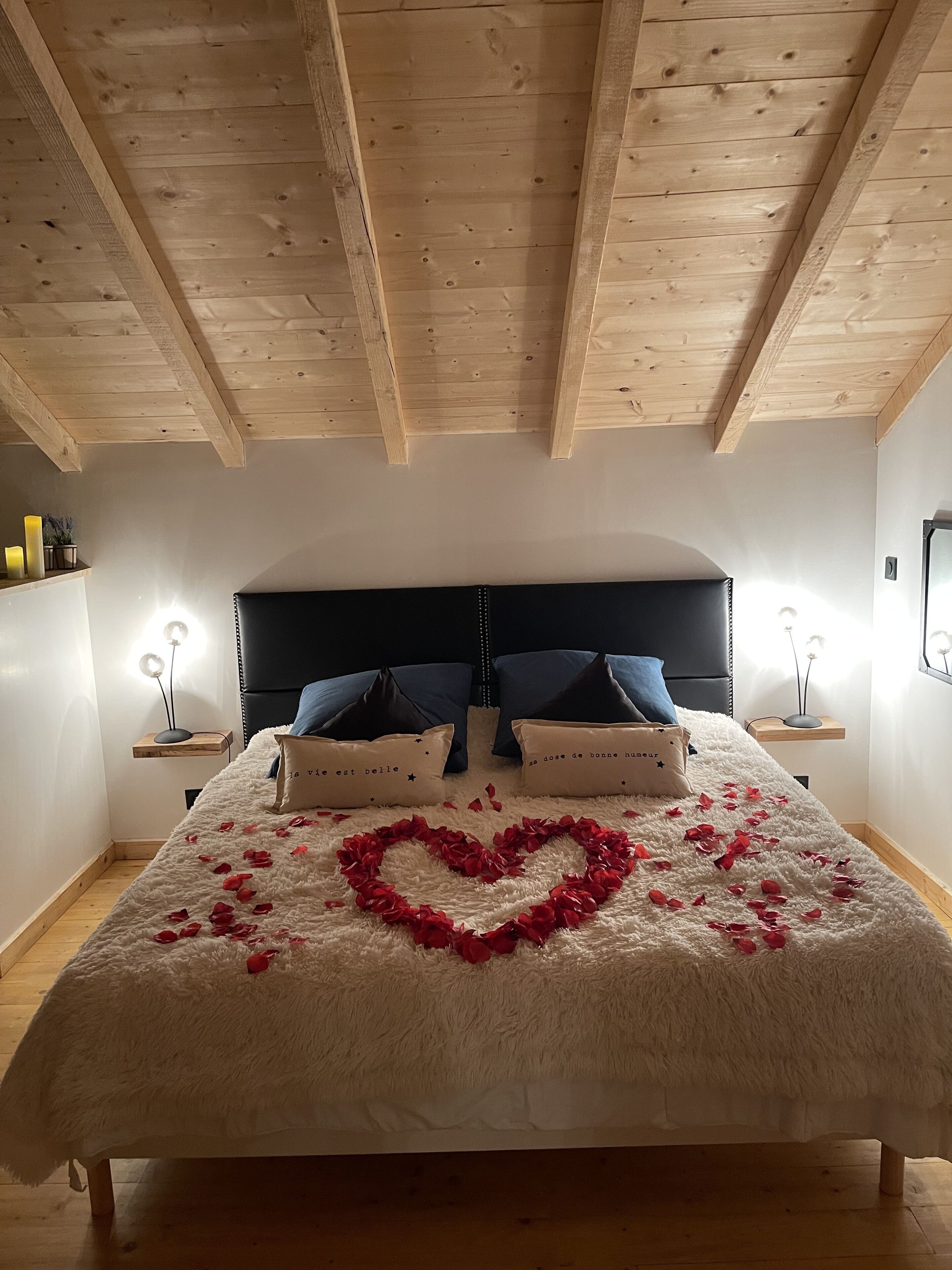 Une chambre avec un plafond en bois, un lit recouvert d’une couette blanche et moelleuse, et une décoration en forme de cœur faite de pétales de roses rouges.