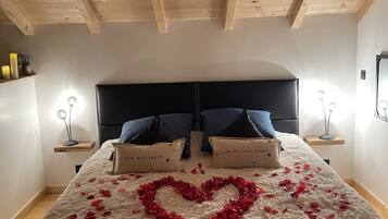 Une chambre avec un plafond en bois, un lit recouvert d’une couette blanche et moelleuse, et une décoration en forme de cœur faite de pétales de roses rouges.