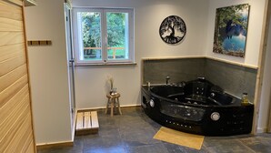 Une salle de bain moderne avec une grande baignoire à remous noire, une fenêtre donnant sur un paysage verdoyant, un tabouret en bois agrémenté d'objets décoratifs et une œuvre d'art fixée au mur.