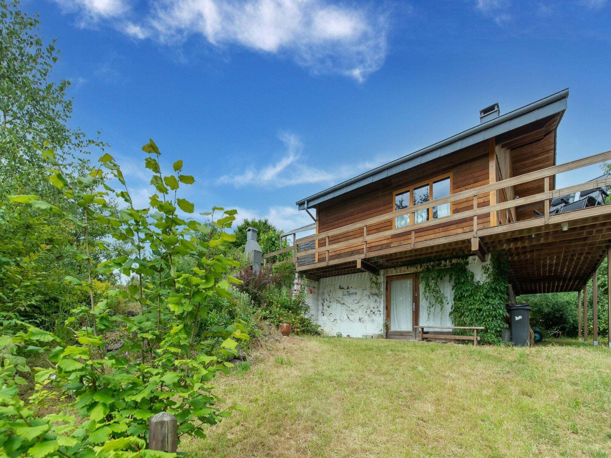 A two-story wooden house with a balcony, surrounded by greenery and a clear sky.