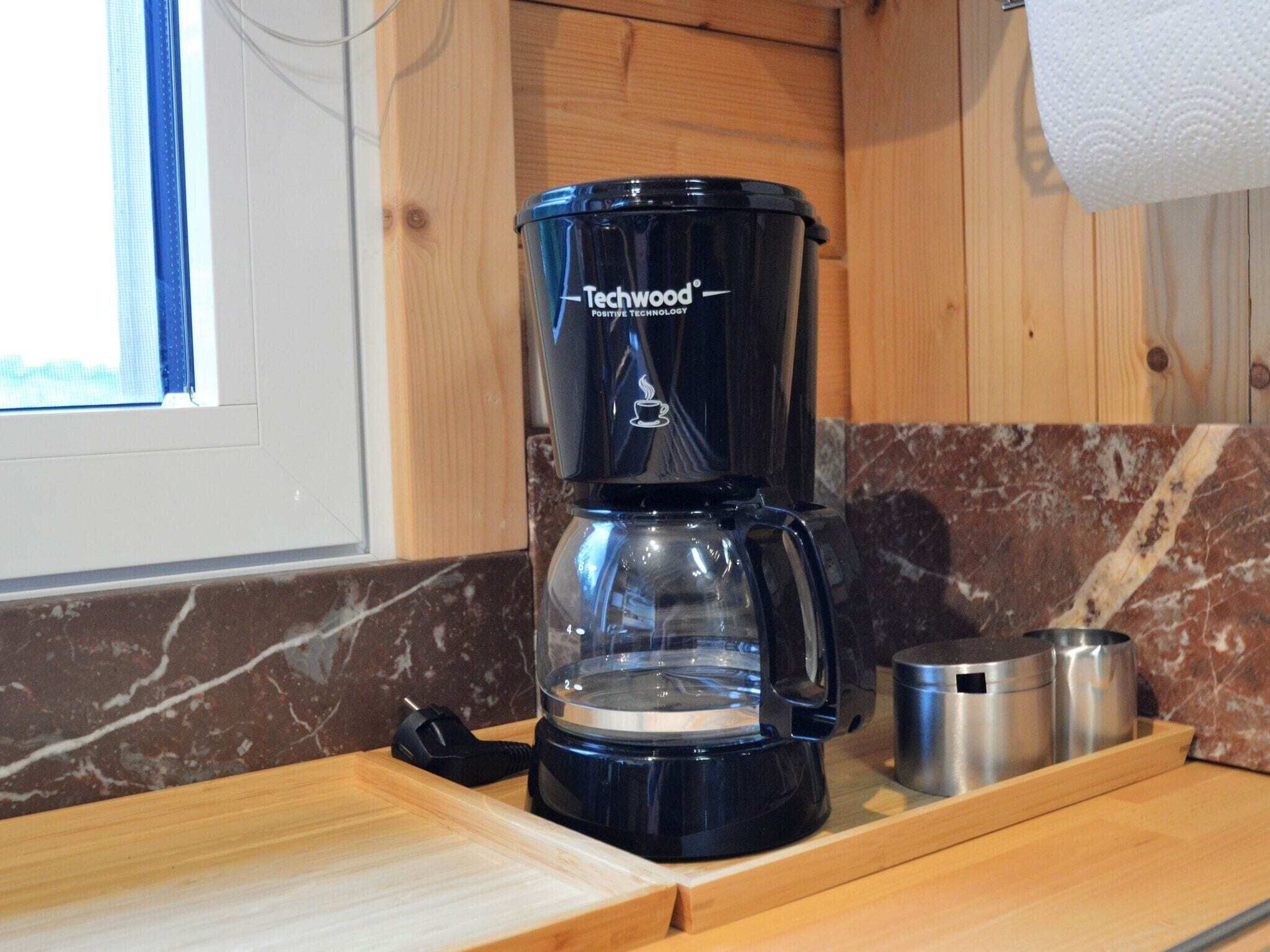 A black coffee maker with a clear carafe, placed on a wooden countertop in a kitchen with a window and marble backsplash.