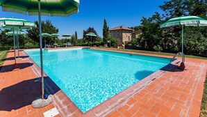 Una piscina rectangular con borde de azulejos, rodeada por un patio de ladrillo. Varias sombrillas verdes ofrecen sombra.
