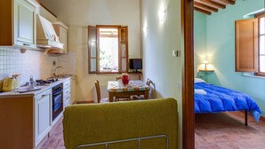 Cocina compacta con zona de comedor, un pequeño televisor y un dormitorio con colcha azul.