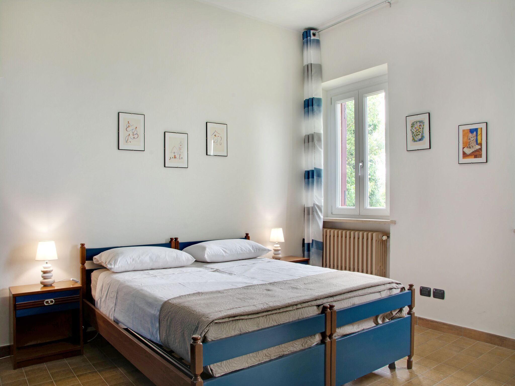 Een slaapkamer met een bed, twee nachtkastjes, een raam met gordijnen en ingelijste foto's aan de muur.