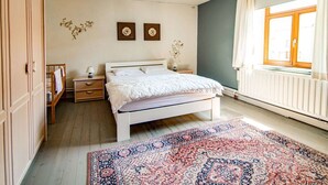 Un dormitorio con cama, mesitas de noche, cómoda, ventana con cortinas y radiador.