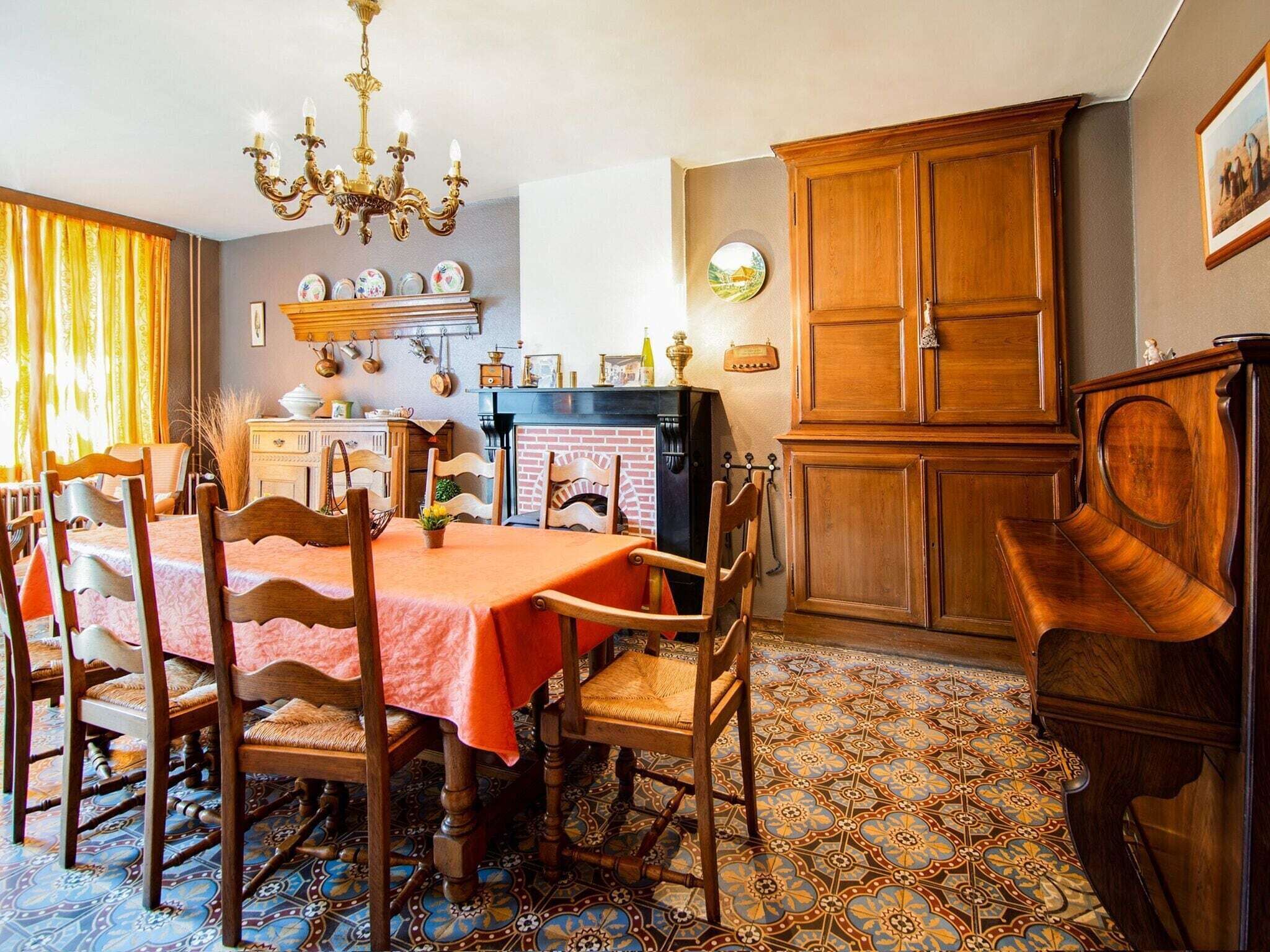 Een eetruimte met een tafel voor vier personen, omringd door houten stoelen. Een kroonluchter hangt aan het plafond en een piano staat rechts. De ruimte heeft vloertegels met een patroon en houten kastjes.