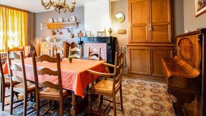 Un comedor con una mesa dispuesta para cuatro personas, rodeada de sillas de madera. Un candelabro cuelga del techo, y un piano se encuentra a la derecha. La habitación tiene baldosas de piso con diseños y armarios de madera.