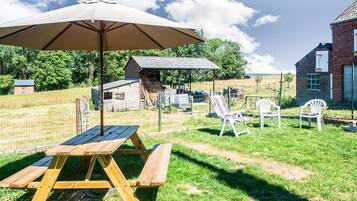 Een picknickopstelling met een houten tafel en bankjes, een parasol, en een omheind gebied met een gebouw op de achtergrond.