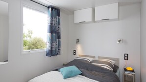 Un dormitorio con cama, ventana con cortinas, mesita de noche con lámpara y estante en la pared.