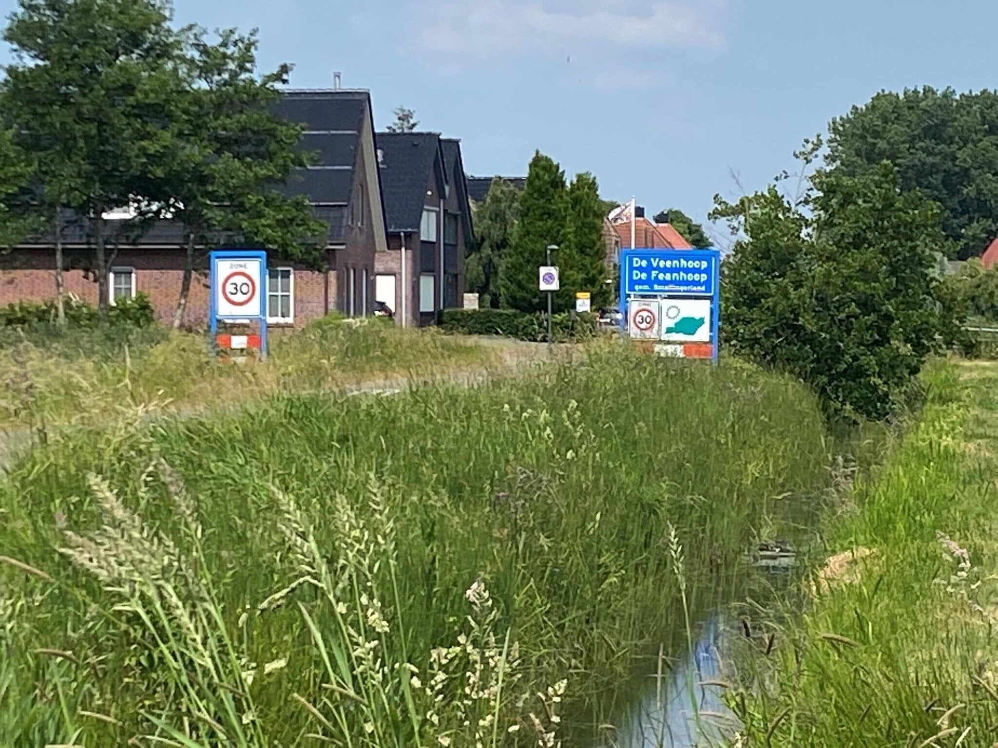 Paisaje rural con un canal, rodeado de hierba alta y árboles. Hay dos señales de tráfico que indican un límite de velocidad de 30 km/h.