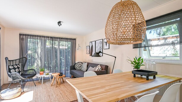 A modern living room with a wooden dining table, white chairs, a grey sofa, and a wicker hanging light fixture. - Modern chalet in Voorthuizen near the forest (Voorthuizen)