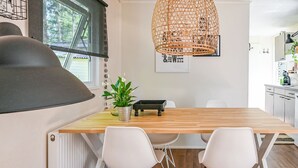 A modern kitchen with a wooden table, white chairs, a hanging rattan light fixture, and a window with a view of greenery. - Modern chalet in Voorthuizen near the forest (Voorthuizen)