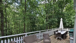 A covered deck with wicker furniture, a table, and chairs, surrounded by a white picket fence and dense trees. - Stylish Chalet with Room (Durbuy)