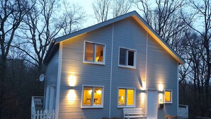 A two-story house with a white picket fence, multiple lit windows, and a bench in front. - Stylish Chalet with Room (Durbuy)
