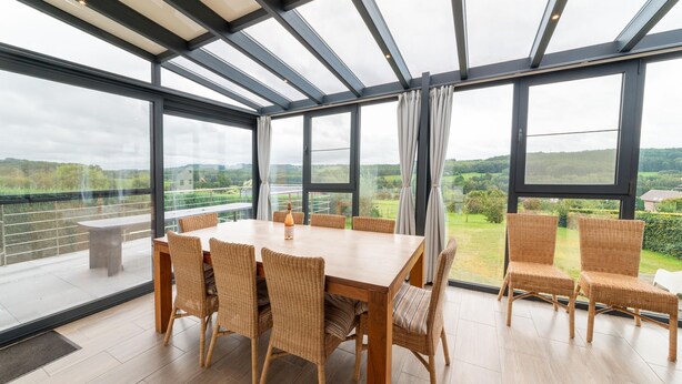 Un balcón espacioso con una mesa de madera y sillas de mimbre, ventanas amplias con cortinas y vistas a zonas verdes y colinas.
