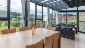 Un comedor con mesa de madera y sillas de mimbre, un sofá y ventanas amplias con vistas al campo.