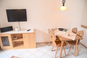A room with a TV mounted on the wall, a wooden desk with a computer, a table with chairs, and a hanging light fixture. - Ferienwohnung in Bayrischzell (Bayrischzell)