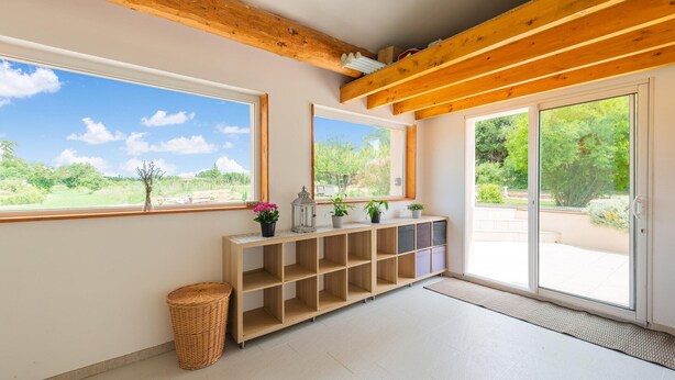 A room with a wooden beam ceiling, large windows with a view of greenery, a built-in shelf with potted plants, and a sliding glass door leading outside.