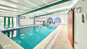 Een groot binnenzwembad met helder water, omringd door een betegelde vloer en een hoog plafond met groene balken.