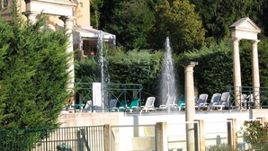 Un'area piscina con fontana, poltrone prendisole e un edificio con colonne sullo sfondo.