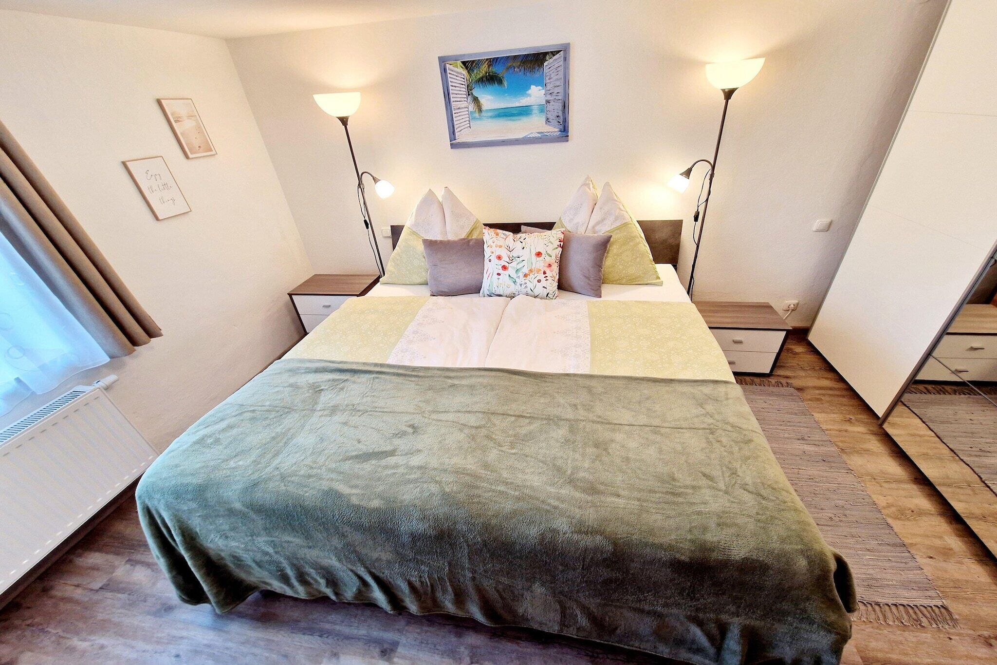 A neatly made bed with a green blanket, two bedside lamps, a wall-mounted painting, and a window with curtains.