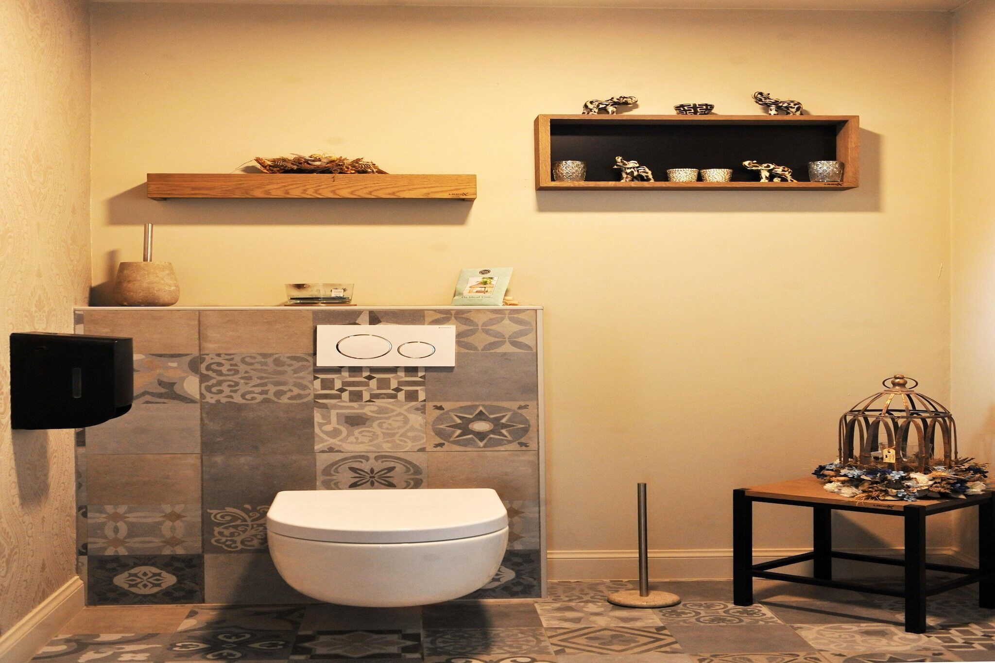 Une salle de bain avec des toilettes, des carreaux décoratifs, une étagère avec des objets décoratifs et une petite table sur laquelle est posée une cage à oiseaux.