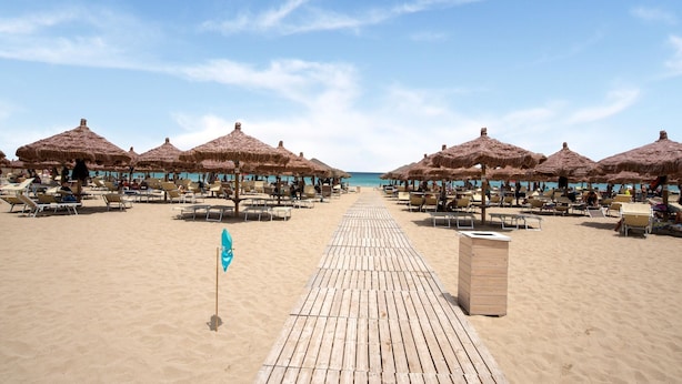 Ein Strand mit Reihen von Liegestühlen und Reetdach-Sonnenschirmen, einem Holzsteg und einem klaren, blauen Himmel.