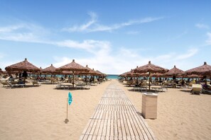 Een strand met rijen ligbedden en rieten parasols, een houten looppad en een heldere blauwe lucht.