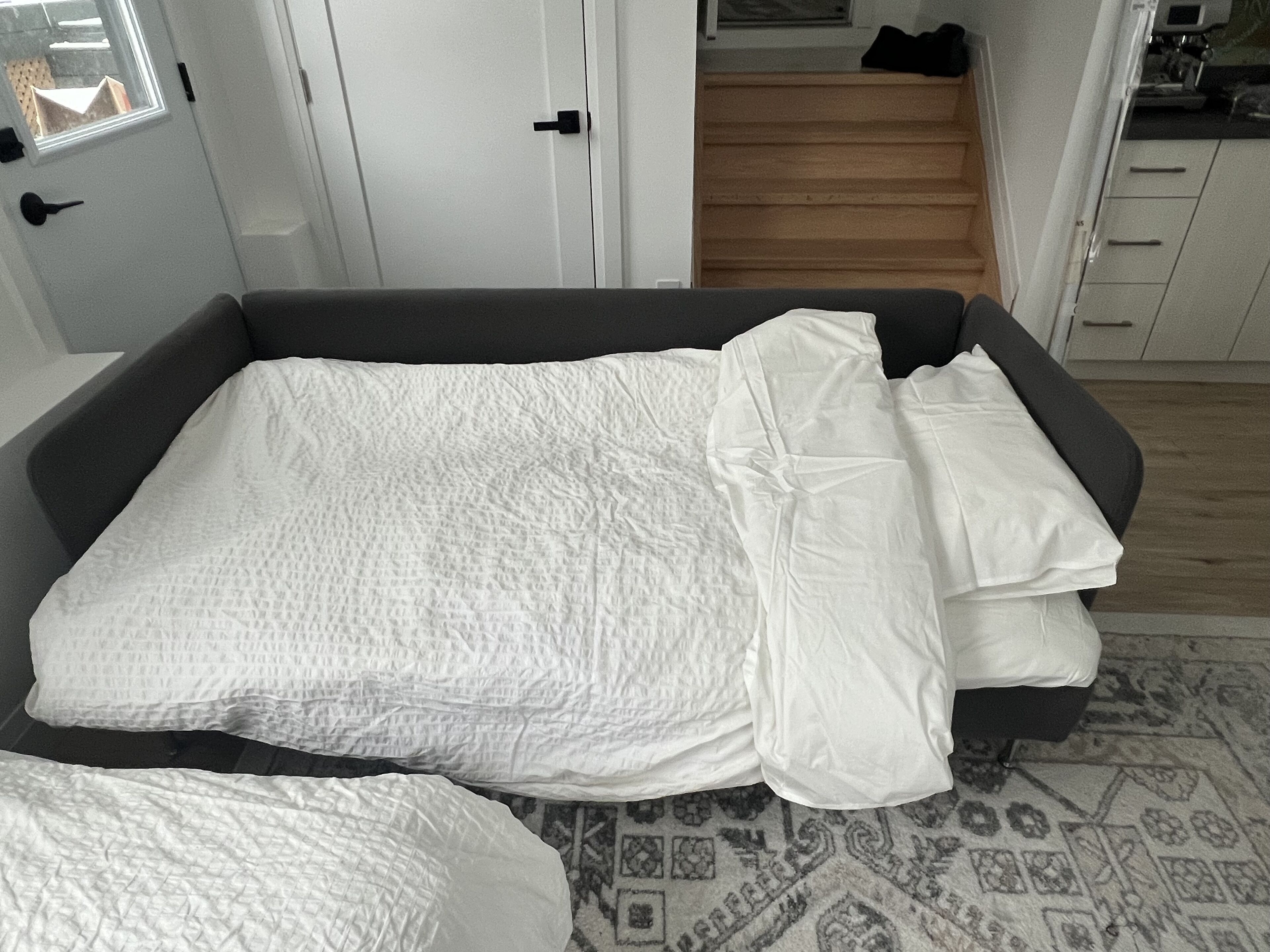 Een net opgemaakt bed met witte beddengoed in een moderne kamer met een grijze bank en een kamerkleed met patroon.