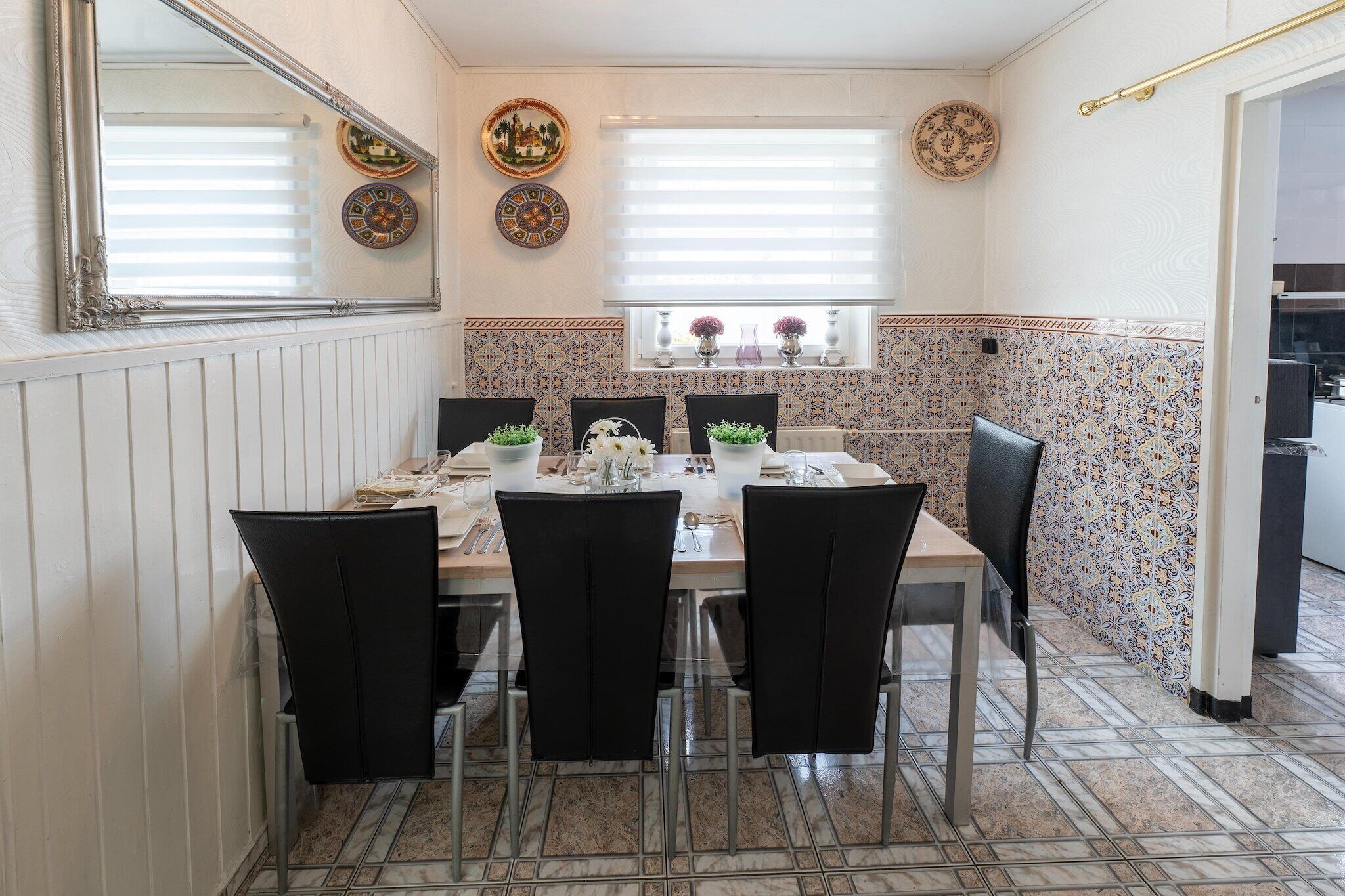 Zona pranzo con tavolo apparecchiato per quattro, sedie nere, piatti decorativi appesi al muro e una finestra con persiane.
