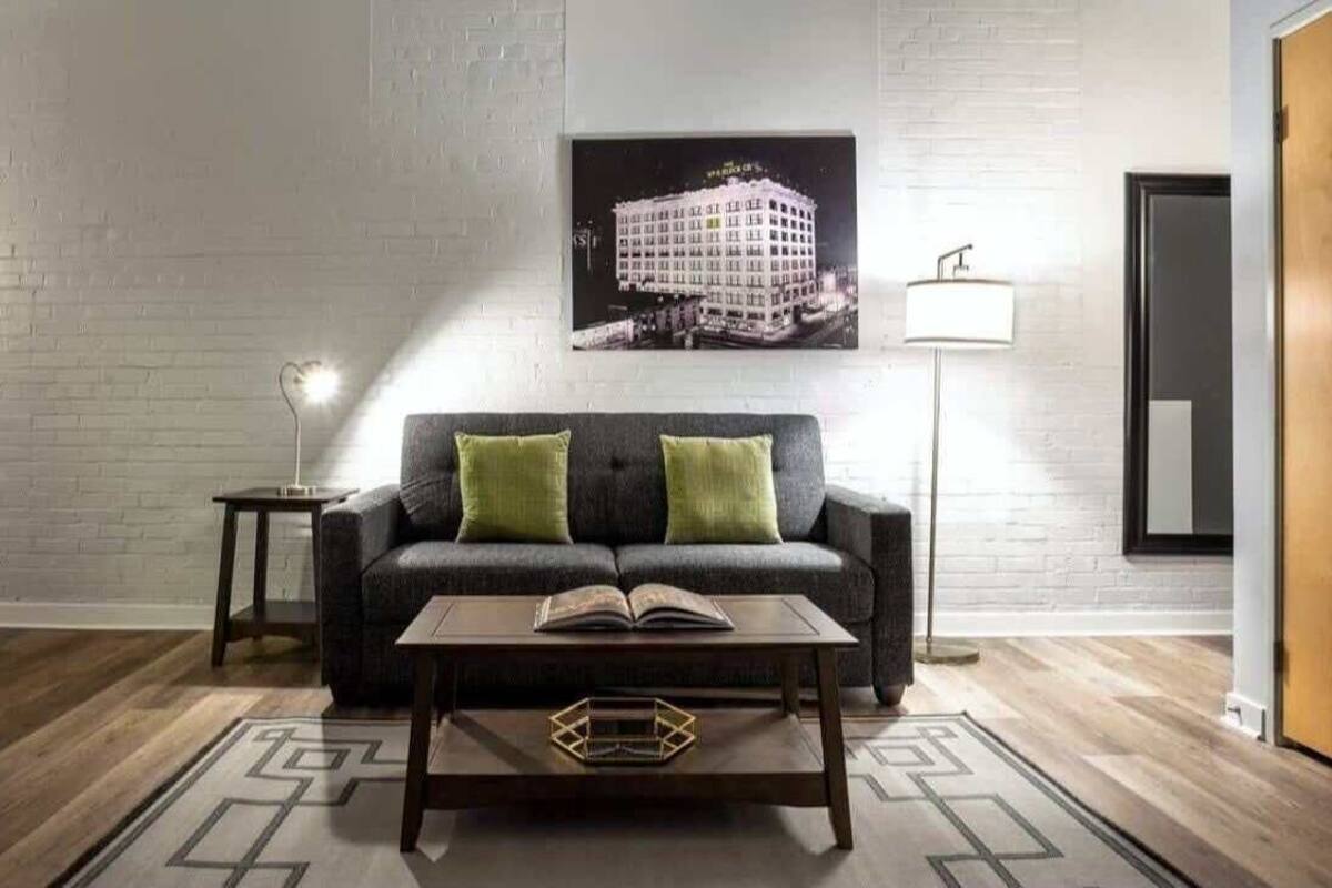 A modern living room with a dark grey sofa, two green cushions, a coffee table with an open book, a framed picture on the wall, and a floor lamp.