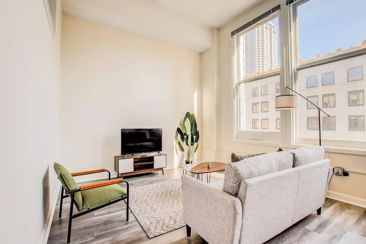 A modern living room with a sofa, a chair, a TV, a plant, and a window with a view of buildings.