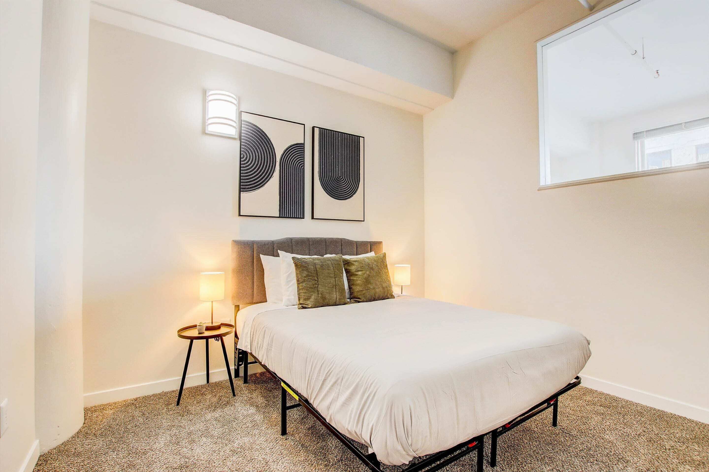 A neatly made bed with a light-colored duvet, a small bedside table with a lamp, and two abstract wall art pieces above the bed.