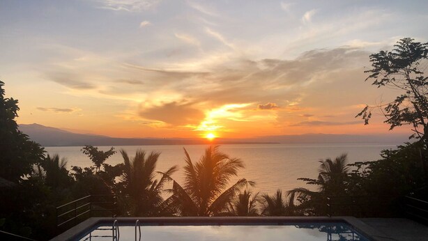 Une piscine offrant une vue sur le coucher de soleil, entourée de palmiers et avec des montagnes en toile de fond.