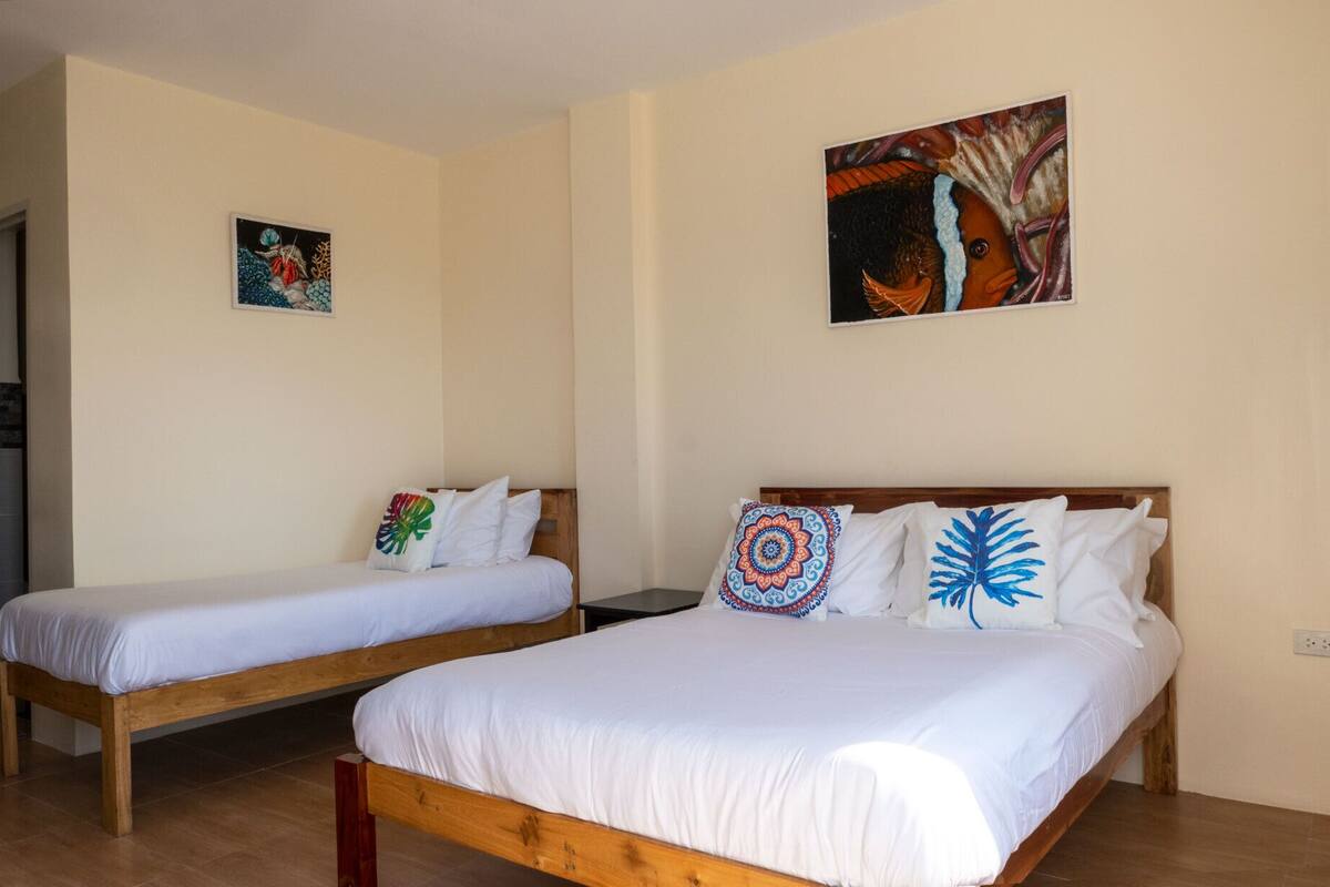 A hotel room with two single beds, each with white bedding and colorful decorative pillows. There are two paintings on the walls, one depicting a tropical scene and the other an abstract design.
