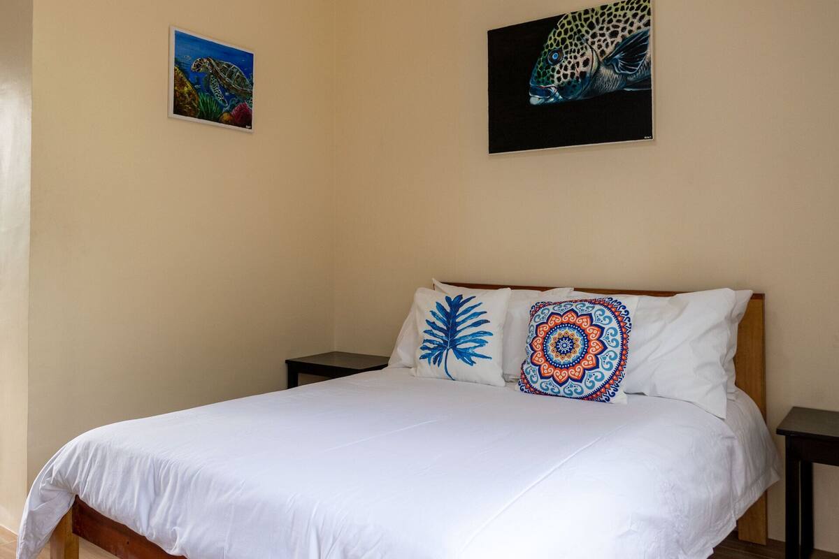 A bed with white bedding, two pillows with blue and colorful patterns, and two framed pictures on the wall.