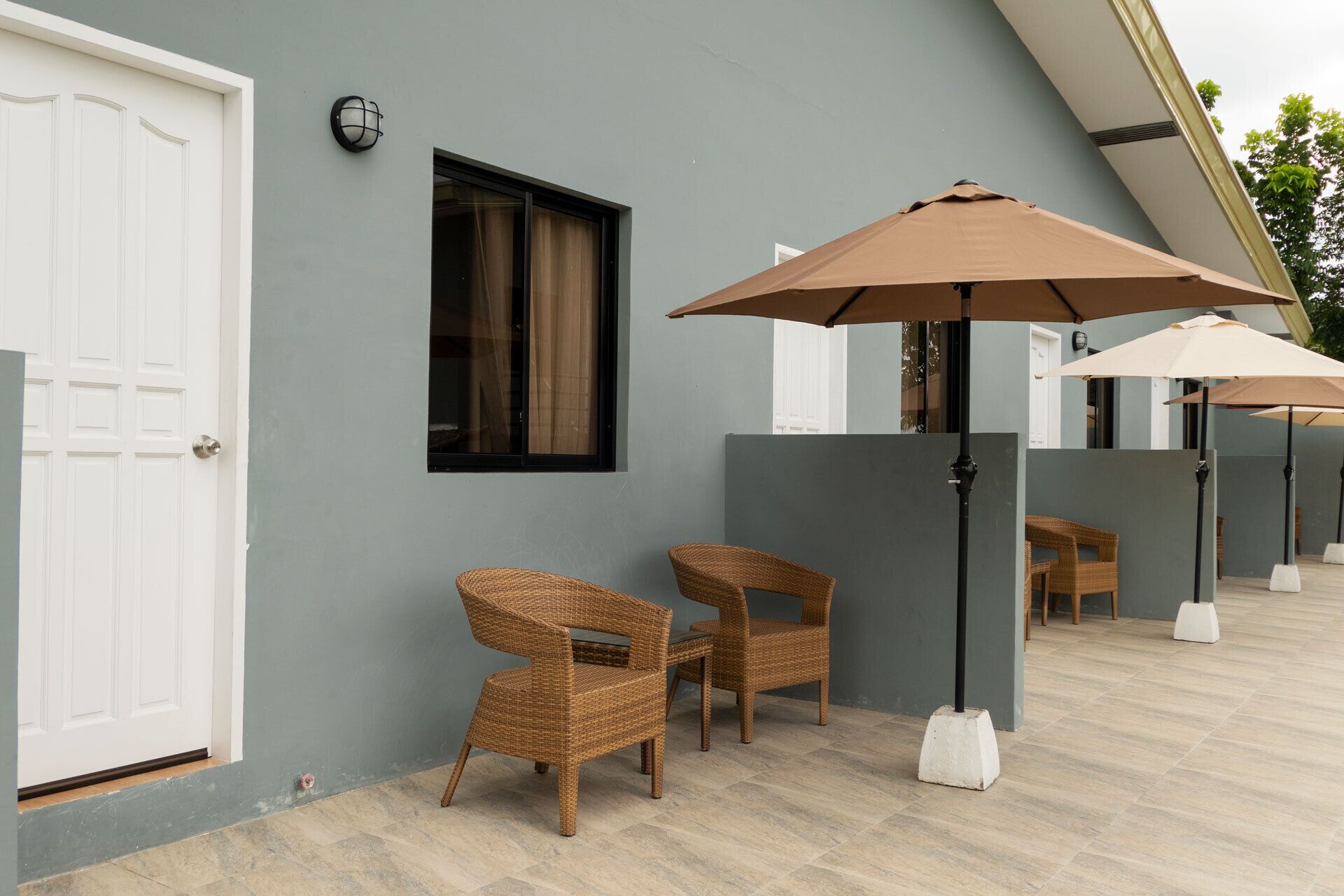A modern outdoor seating area with wicker chairs and umbrellas, a white door, and a window with curtains.