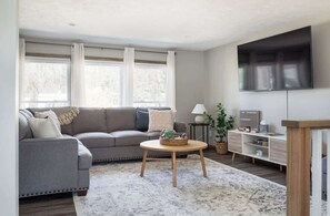 Una sala de estar con un sofá gris, una mesa redonda de centro, un televisor montado en la pared y una planta sobre una mesita auxiliar.