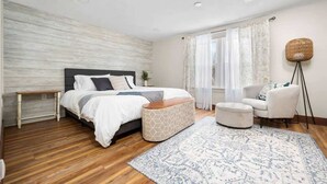 Un dormitorio con cama, mesita de noche, reposapiés, silla y lámpara de pie.