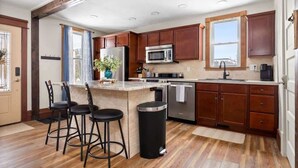 Una cocina moderna con armarios de madera, un refrigerador de acero inoxidable, un microondas y un lavamanos. Hay tres taburetes de bar y un cubo de basura.