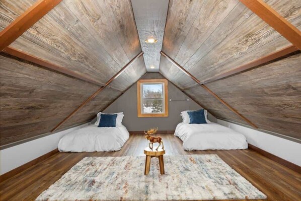 Une chambre située dans un espace de type « loft », avec un plafond en pente, des poutres en bois, deux lits avec une literie blanche, un tapis et une sculpture de cerf en bois.