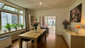 Ein modernes Wohnzimmer mit einem Holzesstisch, Stühlen, einer Couch und einem Couchtisch. Auf dem Fensterbrett stehen Blumentöpfe und an der Wand hängt ein Gemälde.