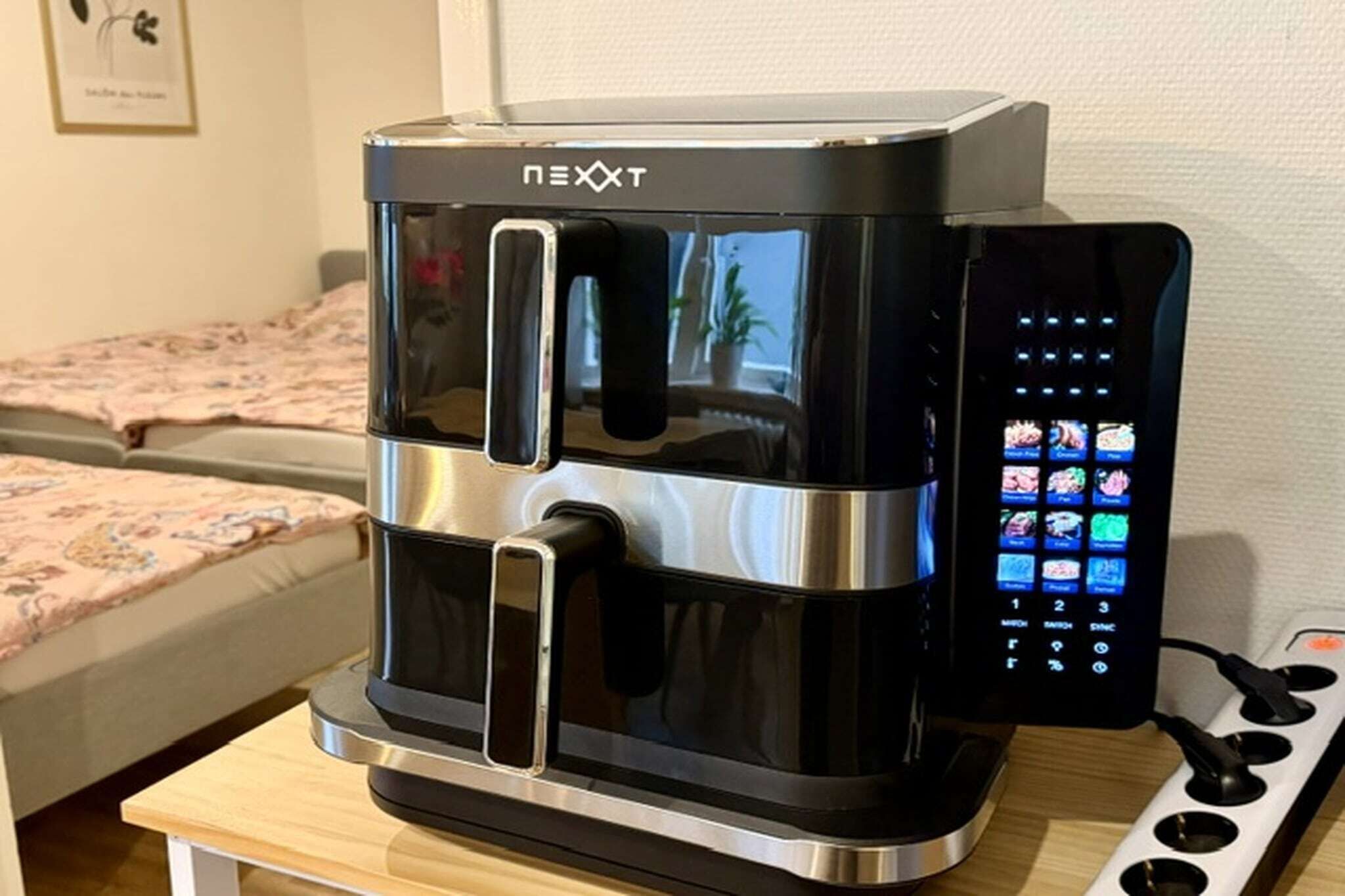 A NEXXT coffee machine with a digital interface, placed on a wooden table in a room with a bed in the background.