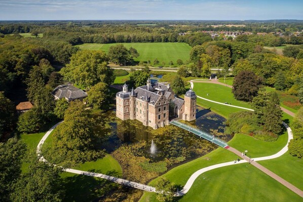 A large, historic castle with a moat, surrounded by lush greenery and a well-maintained lawn.