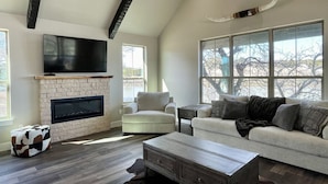 A spacious living room with a stone fireplace, a flat-screen TV, and a large window offering a view of trees.