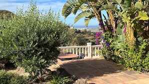 A patio with a table and chairs, surrounded by lush greenery and a view of the sea.