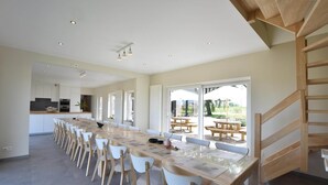 A spacious dining area with a long table and white chairs, a kitchen in the background, and large windows offering a view of the outdoors.
